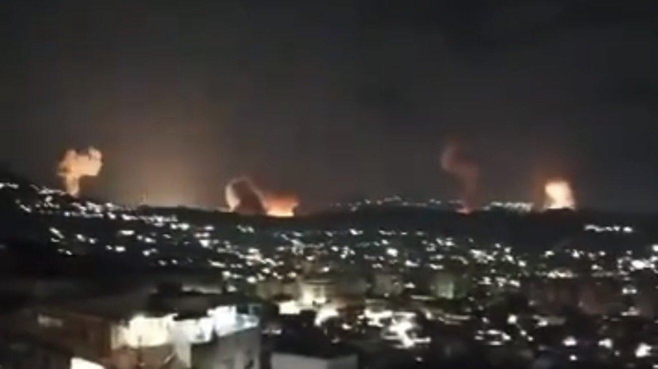 Nighttime view of a city skyline illuminated by distant fires and explosions on the hills, showcasing a dramatic and intense scene.