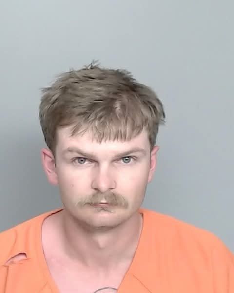 Mugshot of a man with a mustache wearing an orange prison jumpsuit, looking directly at the camera against a neutral background.