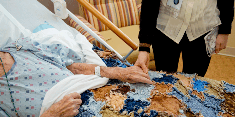 A hospice patient is in bed with her hand being held by a hospice caregiver.