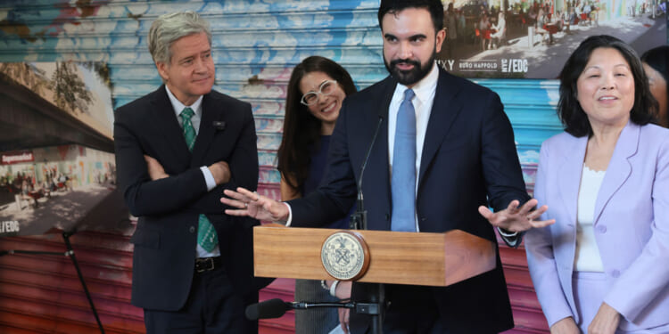 New York Mayor Zohran Mamdani stands at a little podium with his arms wide in front of a grafitti-art filled wall. Three associates surround him.