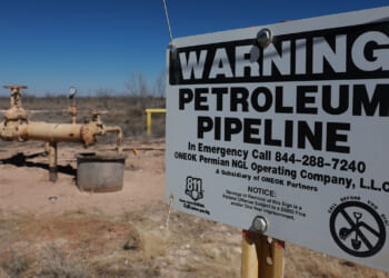 Yellow oil pipelines in a barren West Texas landscape. In the foreground, a sign reading "Warning Petroleum Pipeline"