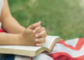 Close-up of young person with flag and Bible on their lap in outdoor setting.
