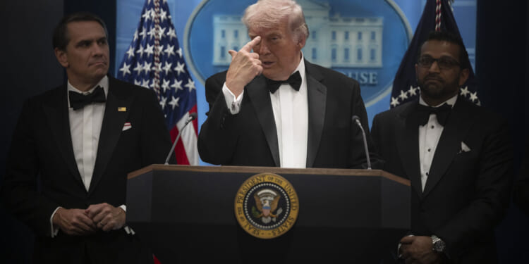 Donald Trump in a tux points to his forehead during a press briefing. He's flanked by Kash Patel and Todd Blanche.