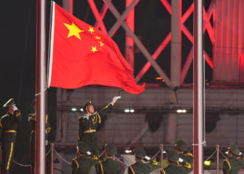 Chinese soldier points to large Chinese flag as row of Chinese soldiers salute.