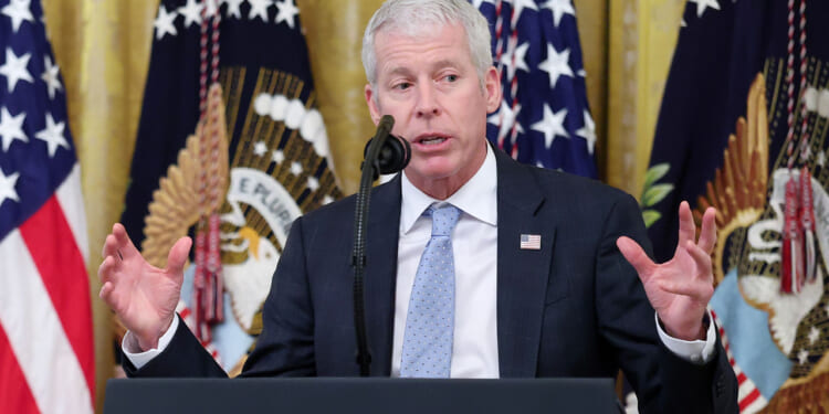 Energy Secretary Chris Wright holds his hands up while speaking behind a podium with flags behind him.