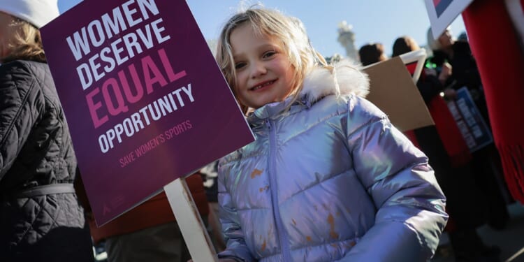 A young girl protests with a sign that says "Women deserve equal opportunity. Save women's sports."