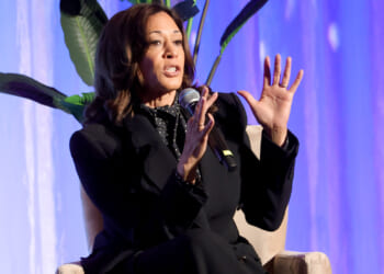 A seated Kamala Harris raises her hands as she makes a point during a talk.