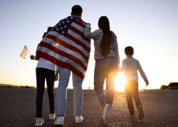 A mother and father walk with their two kids toward a sunset, a flag draped on the father's back and a flag in the hand of the taller sibling.