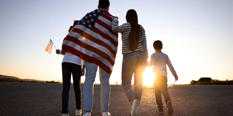 A mother and father walk with their two kids toward a sunset, a flag draped on the father's back and a flag in the hand of the taller sibling.