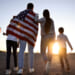 A mother and father walk with their two kids toward a sunset, a flag draped on the father's back and a flag in the hand of the taller sibling.