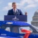 Transportation Secretary Sean Duffy speaking at a podium in front of a car
