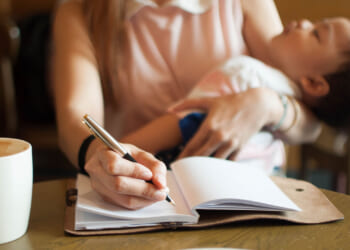 A mother writes in a notebook while holding her sleeping child at a table.
