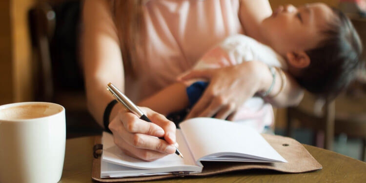 A mother writes in a notebook while holding her sleeping child at a table.