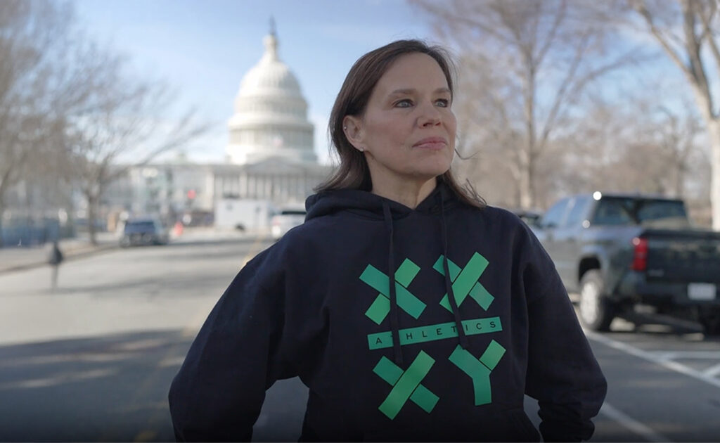 How a Pro-Reality Sports Brand Aims to Compete With Transgender-Aligned Nike 2 A woman in a black hoodie with green text stands on a street with the U.S. Capitol building visible in the background on a clear day.