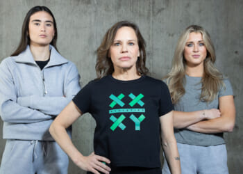 Three women stand in front of a concrete wall. The woman in the center, Jennifer Sey, wears a black XX-XY Athletics t-shirt, while the other two, Paula Scanlan and Riley Gaines in gray outfits, stand on either side with arms crossed. All face the camera with serious expressions.