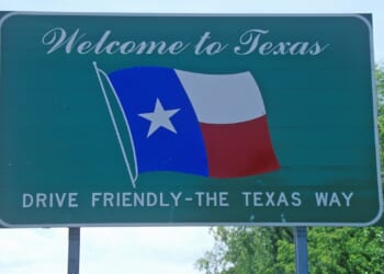 Green roadside sign with Welcome to Texas, a waving Texas flag, and the slogan Drive Friendly – The Texas Way against a backdrop of trees and blue sky.