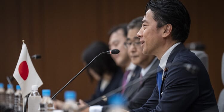 A man in a suit speaks at a conference table with other people, microphones, and a Japanese flag visible. Bottled water is on the table and everyone faces the same direction.