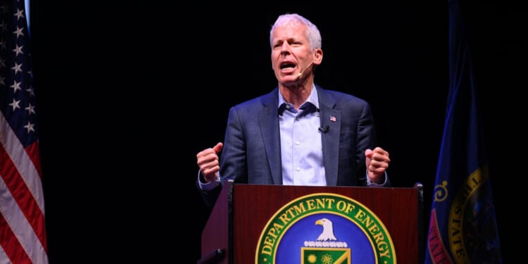 A man speaks passionately at a podium with the U.S. Department of Energy seal, flanked by the American flag and another flag, against a dark background.