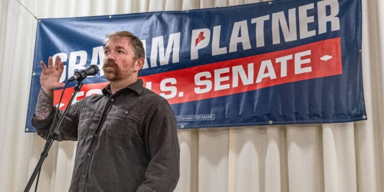 A man stands at a microphone with one hand raised, speaking in front of a campaign banner that reads Graham Platner U.S. Senate against a backdrop of white curtains.