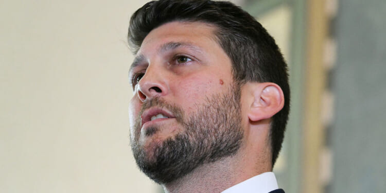 Florida Attorney General James Uthmeier is suing pro-transgender activist groups the American Academy of Pediatrics, the World Professional Association for Transgender Health, and the Endocrine Society. In this picture, Uthmeier with short dark hair and a beard, wearing a suit, looks upward while speaking indoors.