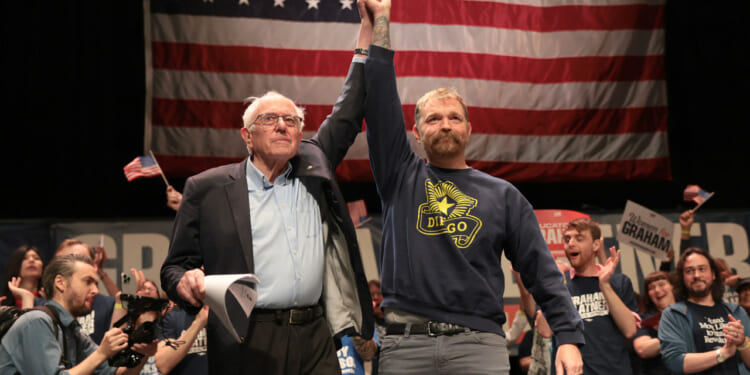An older man in a suit holds up a mans arm on stage in front of a large American flag, with a cheering crowd in the background holding signs and waving flags.