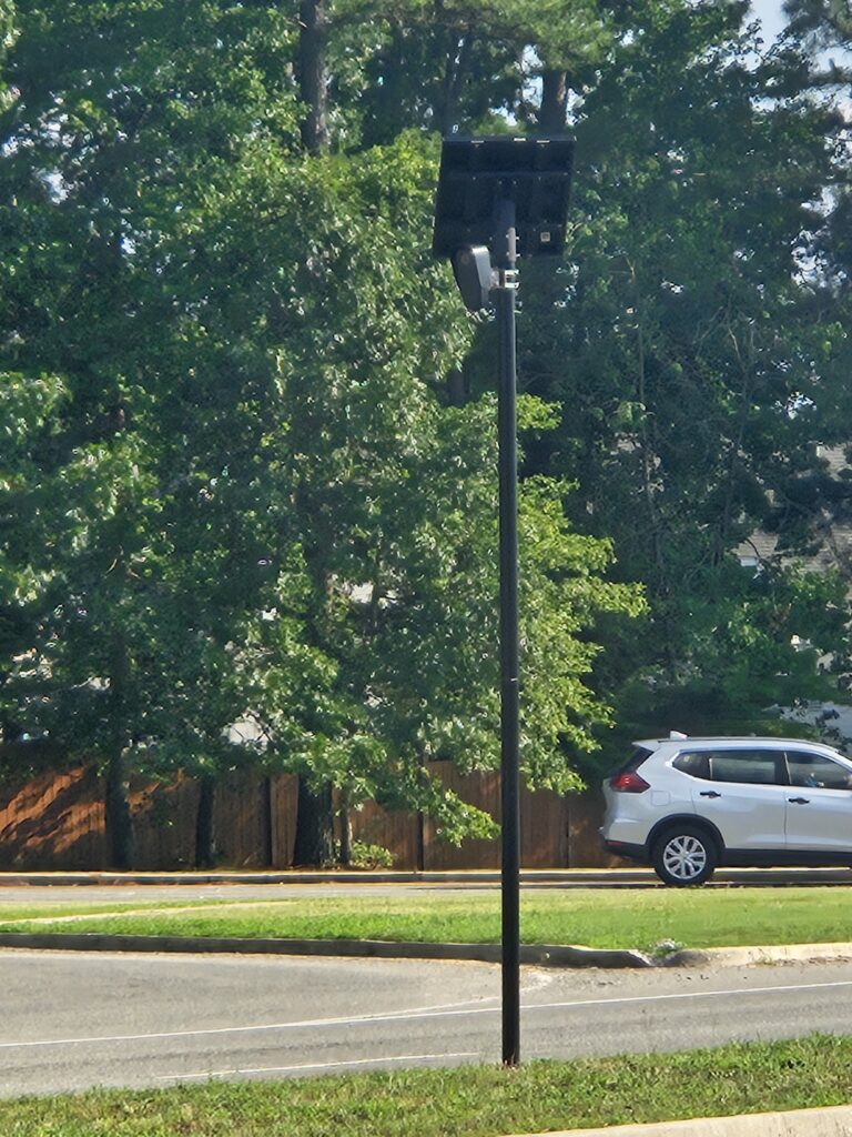 A surveillance camera stands by a road with a white SUV parked nearby. Green trees and a wooden fence are in the background.