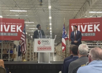Republican nominee for Ohio governor, Vivek Ramaswamy, looks on at his running mate, Senate President Rob McColley as they stand on a stage in a warehouse, with Vivek for Ohio campaign signs and American and Ohio flags behind them.
