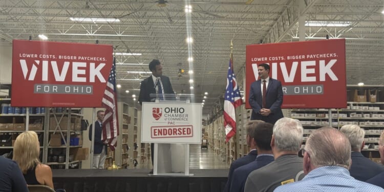 Republican nominee for Ohio governor, Vivek Ramaswamy, looks on at his running mate, Senate President Rob McColley as they stand on a stage in a warehouse, with Vivek for Ohio campaign signs and American and Ohio flags behind them.