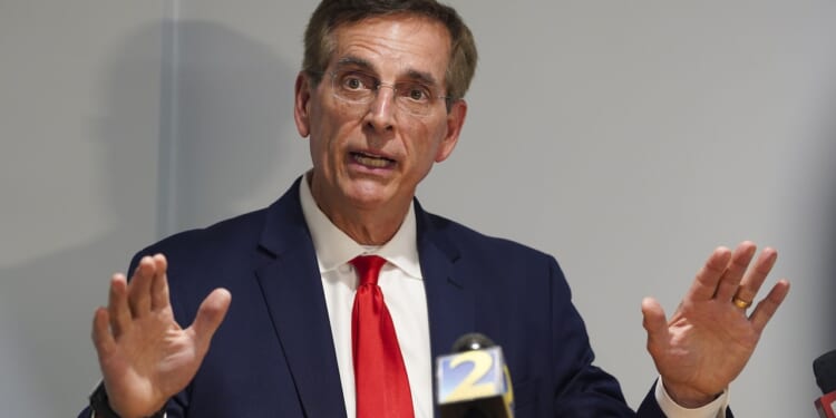 A man in a navy suit and red tie speaks with his hands raised in front of microphones at a press conference.