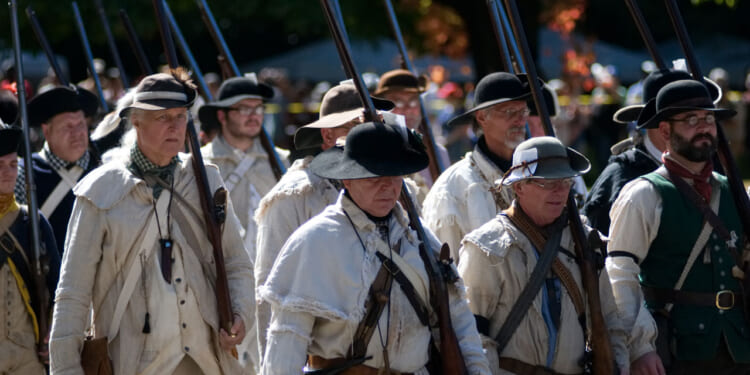 A group of people dressed in historical colonial clothing march outdoors, carrying long rifles upright over their shoulders. Trees with green and orange leaves are visible in the background.