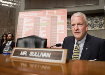 Sen. Dan Sullivan in a suit sits at a desk with a nameplate reading Mr. Sullivan. Behind him is a chart titled Russian & Chinese Incursions Into Alaska in a formal hearing room.
