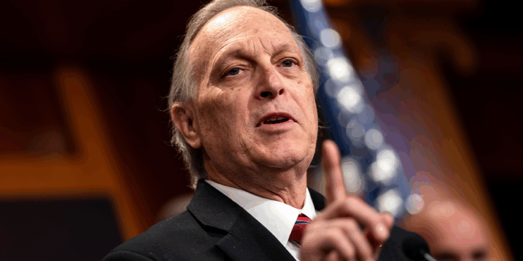 An older man in a suit gestures while speaking at a podium, with an American flag blurred in the background.