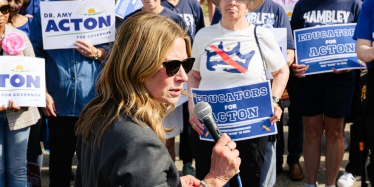 A woman in sunglasses speaks into a microphone at a rally. People behind her hold blue signs reading EDUCATORS FOR ACTION and DR. AMY ACTON FOR GOVERNOR. Some wear shirts supporting Amy Acton and Hambly.