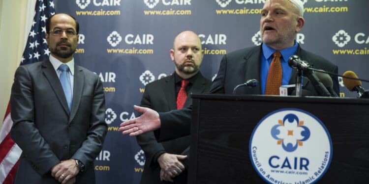 Three men stand at a CAIR podium with microphones. Behind them is a CAIR banner and a U.S. flag. One man is speaking, while another gestures with his hand.