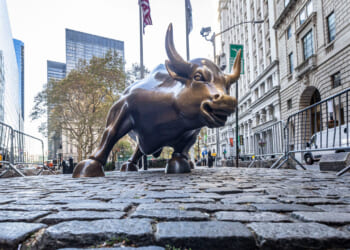 A low-angle view of the bronze Charging Bull statue on cobblestone, surrounded by metal barricades, with tall buildings and trees in the background.