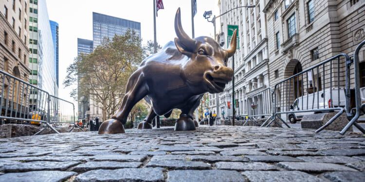 A low-angle view of the bronze Charging Bull statue on cobblestone, surrounded by metal barricades, with tall buildings and trees in the background.