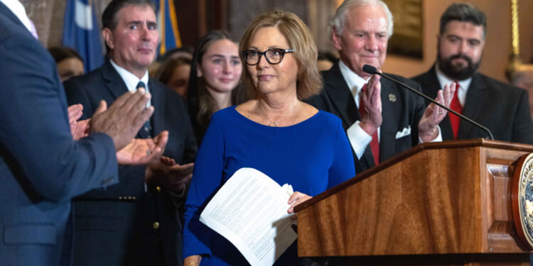 Darline Graham in a blue dress holding papers stands at a podium as people around her applaud. Several people in suits and formal attire are visible, and a flag is in the background.