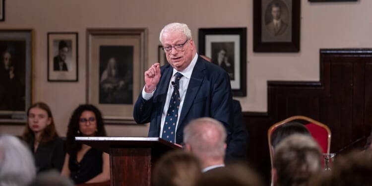 An older man in a suit speaks at a podium in a formal room with framed photos on the wall. Several people, reflecting diversity, are seated and listening attentively in the background.