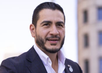 A man with short dark hair and a trimmed beard wearing a dark plaid suit jacket and white shirt stands outdoors, with blurred buildings in the background.