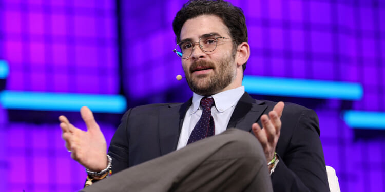 Hasan Piker responded to outrage over his singing the song "We are Charlie Kirk." Pictured: a man in a suit and glasses, wearing a headset microphone, gestures with his hands while speaking on stage against a bright purple and blue geometric background.