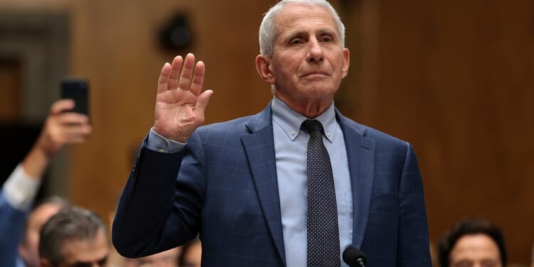 An older man in a blue suit raises his right hand, appearing to take an oath in a formal indoor setting, with people seated and watching in the background.