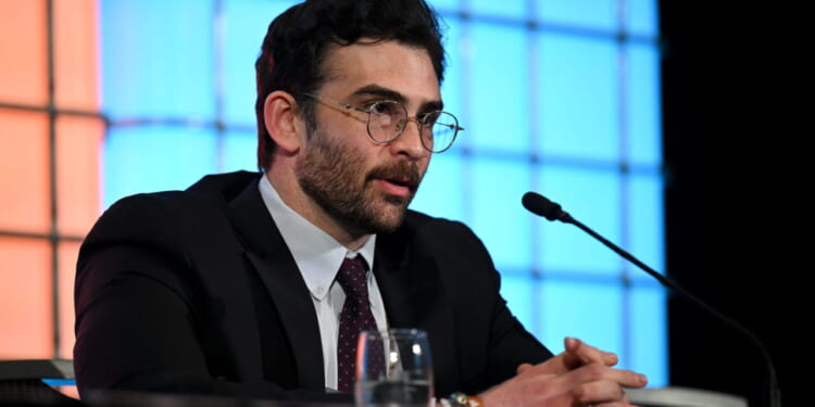 Hasan Piker in a suit and glasses speaks into a microphone at a panel, with his hands clasped on the table. Blue and orange backgrounds are behind him.