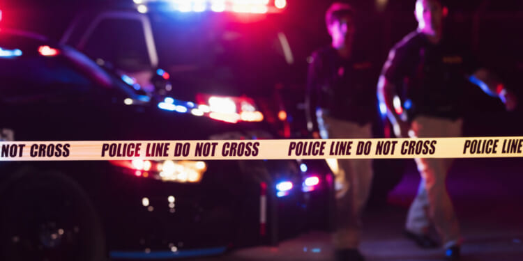 Police tape in the foreground in front of two officers and a police car with flashing lights at night.