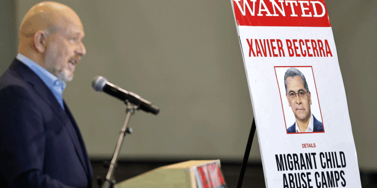 A man speaks at a podium next to a large sign reading WANTED with a photo of Xavier Becerra and the words Migrant Child Abuse Camps in bold letters.