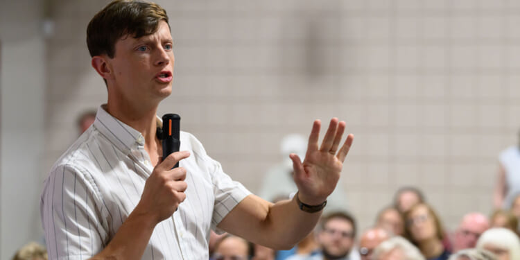 Rob Sand in a striped shirt holds a microphone and speaks, gesturing with his hand, in front of a seated audience in a room with tiled walls.