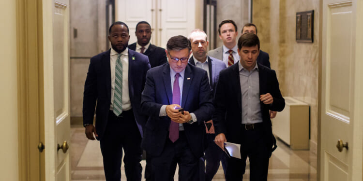 A group of men, including Mike Johnson, in business attire walk down a hallway. The man at the front is looking at his phone, while others follow closely behind. The setting appears to be an official or government building.