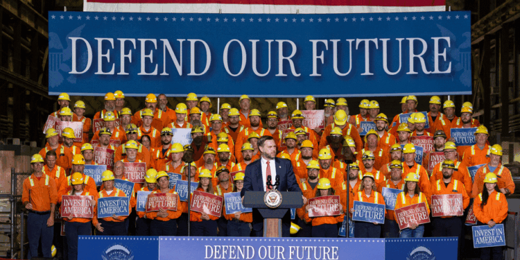 Vice President JD Vance stands at the podium as he addresses a crowd of construction workers in orange gear, with signs reading Defend Our Future and Invest in America. A large banner behind reads DEFEND OUR FUTURE.