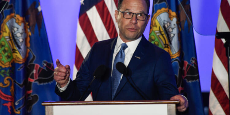 A man in a blue suit, identified as Josh Shapiro, speaks at a podium labeled SHAPIRO, with American and Pennsylvania state flags in the background. He gestures with one hand and looks to the side.
