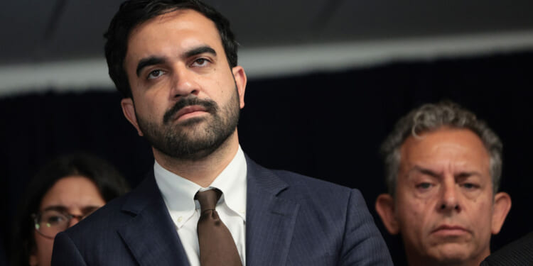 A man in a suit and tie stands in front of a dark background, looking serious. Two other people, also appearing serious, are partially visible beside and behind him.