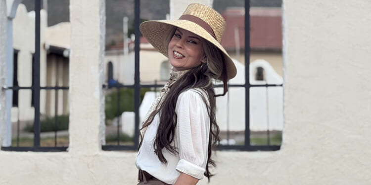 Allison Roberts is suing Portland Public schools over retaliation for her opposition to transgender lessons in school. Pictured: Roberts, a woman in a wide-brimmed straw hat and white blouse smiles while standing outdoors in front of a light-colored wall with iron-barred windows.